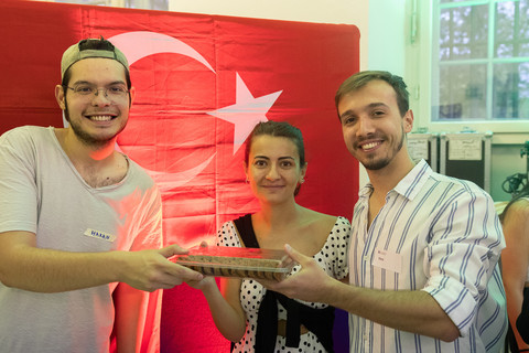 Drei Menschen stehen lächelnd vor einer türkischen Flagge und halten eine türkische Spezialität hoch.